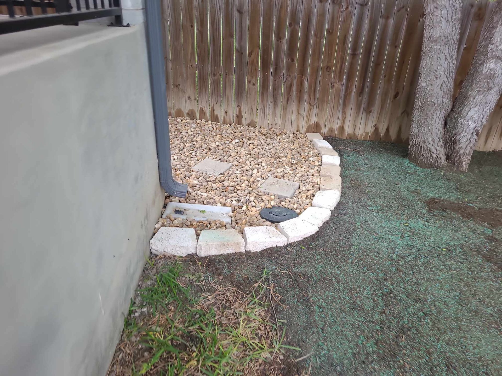 Small backyard corner with a curved stone border, gravel bed, and patchy grass beside a fence and wall.