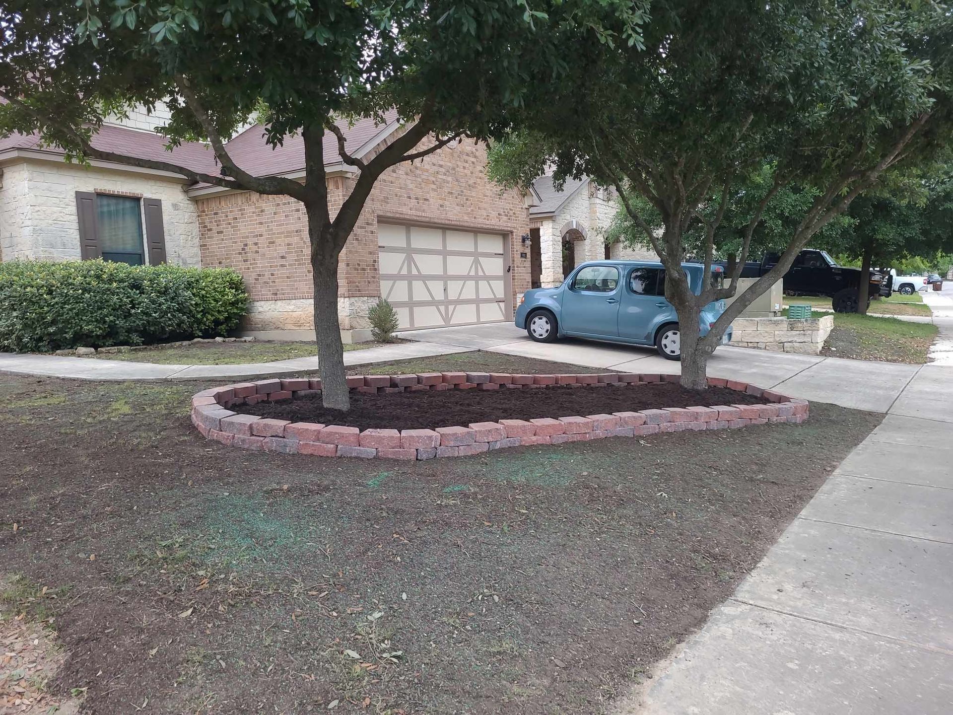 Front yard with two trees, a blue car, and a house with a garage beside a sidewalk.