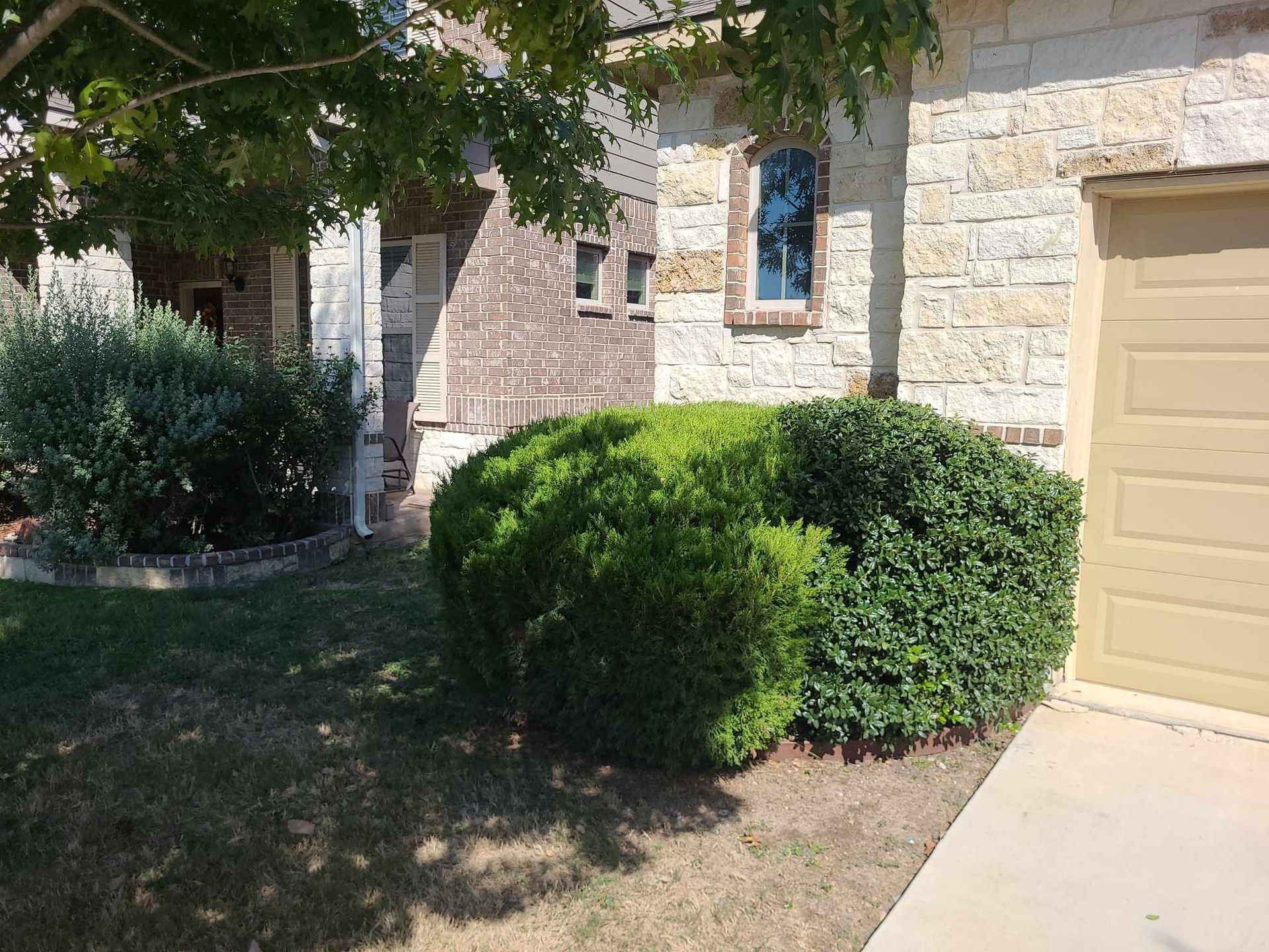 Stone house exterior with tan garage door, trimmed green bushes, and shaded front yard
