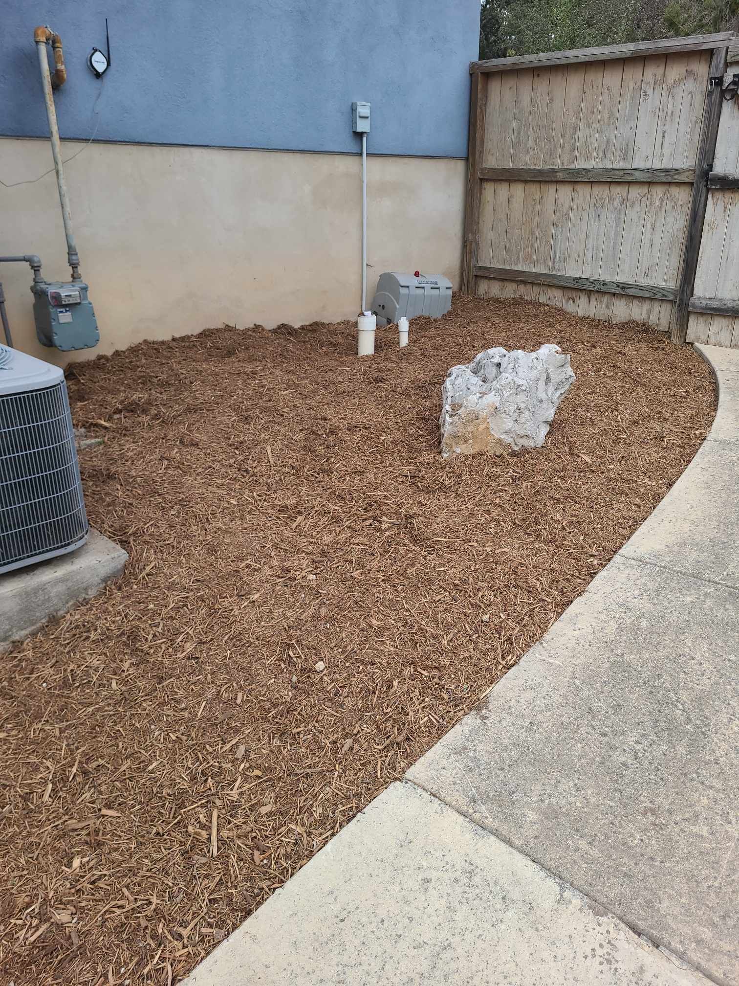 Backyard corner with brown mulch, utility pipes, a fence, and a concrete walkway.