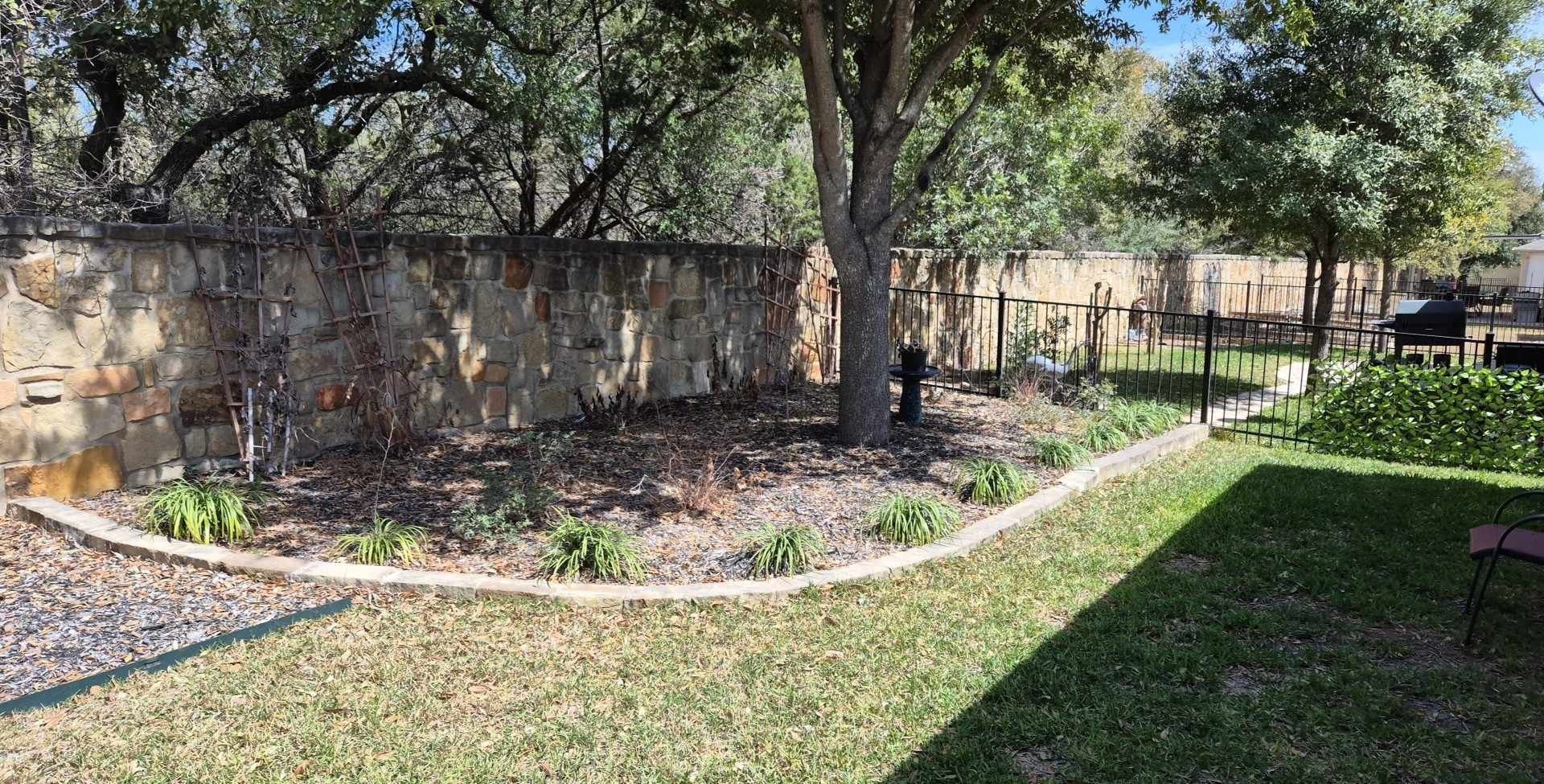Backyard with grass, trees, stone edging, and a shaded garden bed along a weathered wall