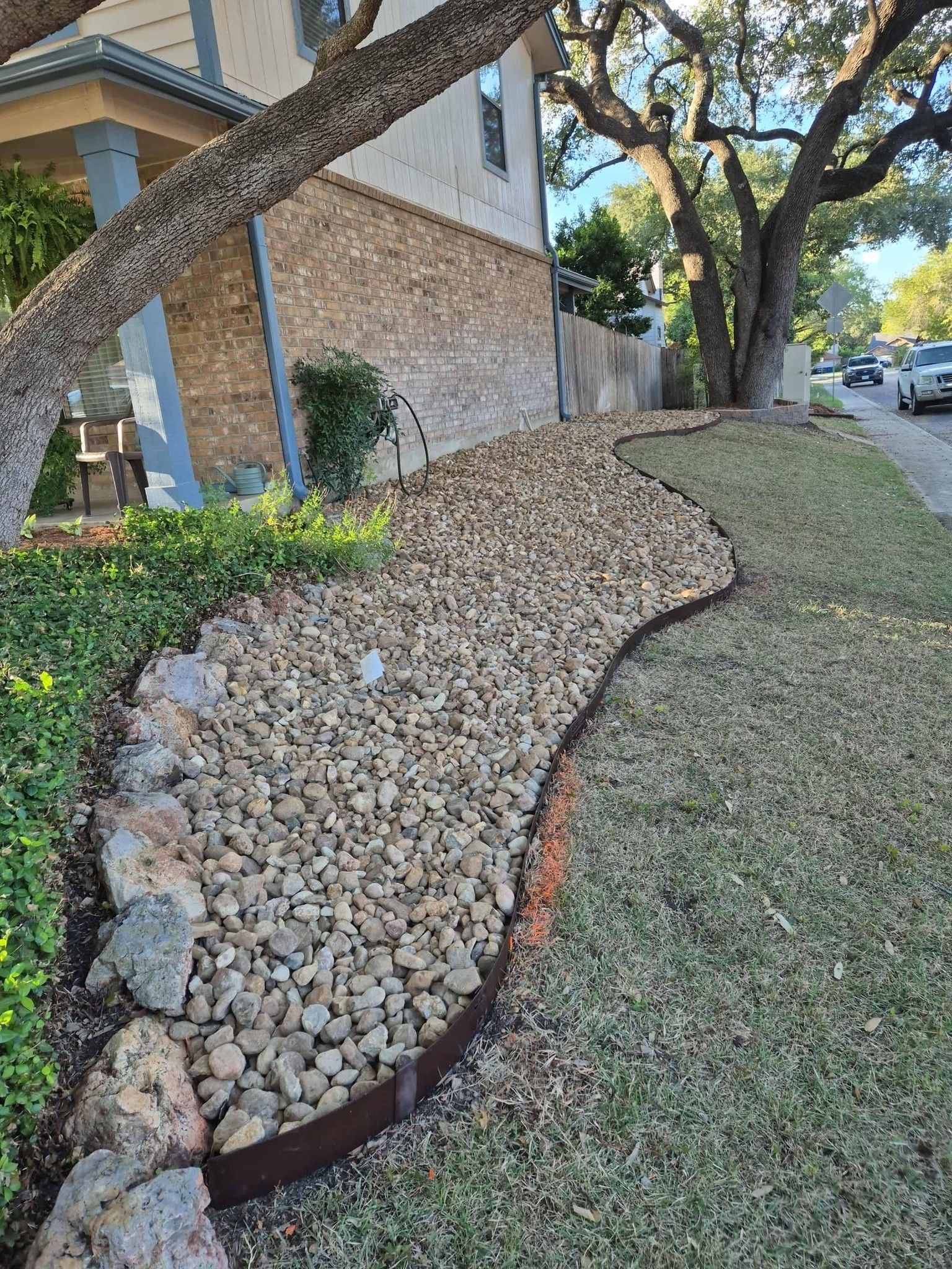 Rock-lined landscaped yard with sloped mulch border beside a brick house and sidewalk.