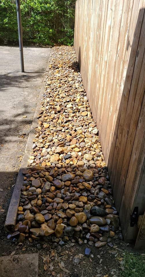 Narrow gravel drainage strip between a wooden fence and paved path, with scattered stones and a small curb.