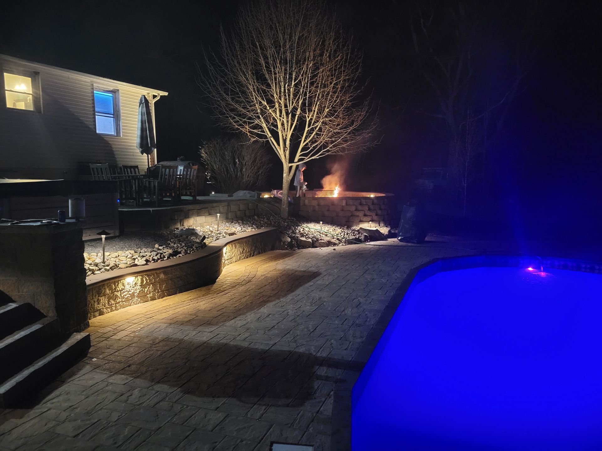 Night view of backyard with lit pool, fire pit, and tree, beside a house with some lights on.