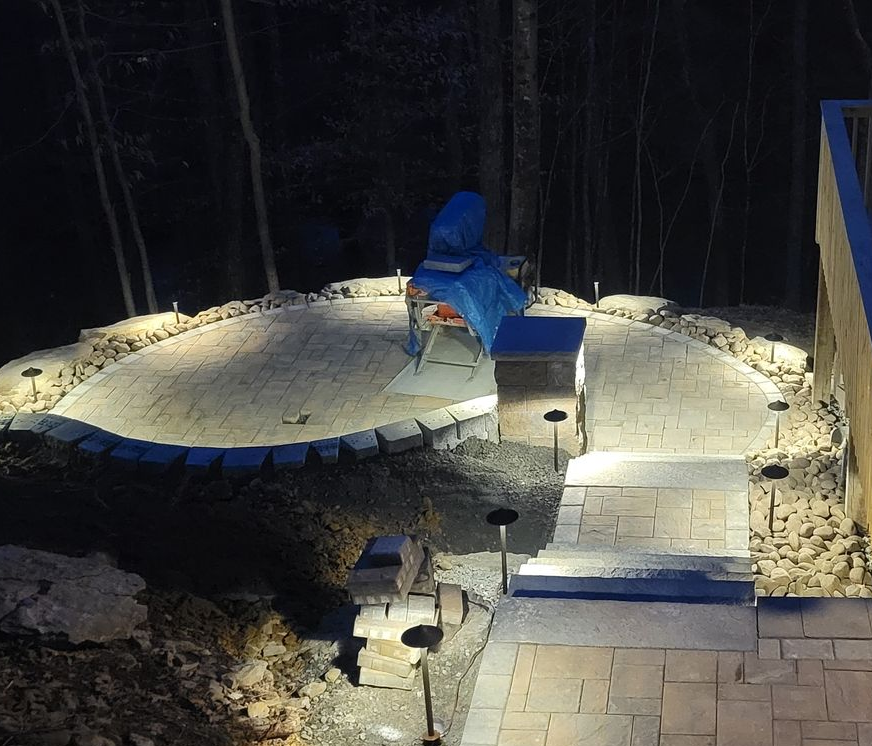 Patio with a fire pit and seating, paved with gray blocks; a deck and landscaping are in the background.