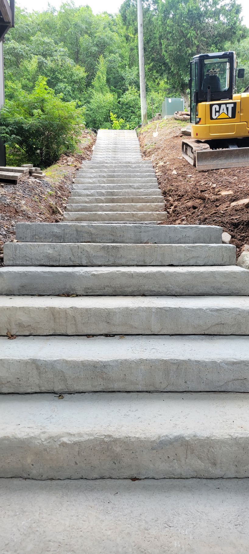 Concrete steps leading uphill towards green trees with a yellow CAT excavator on the right.