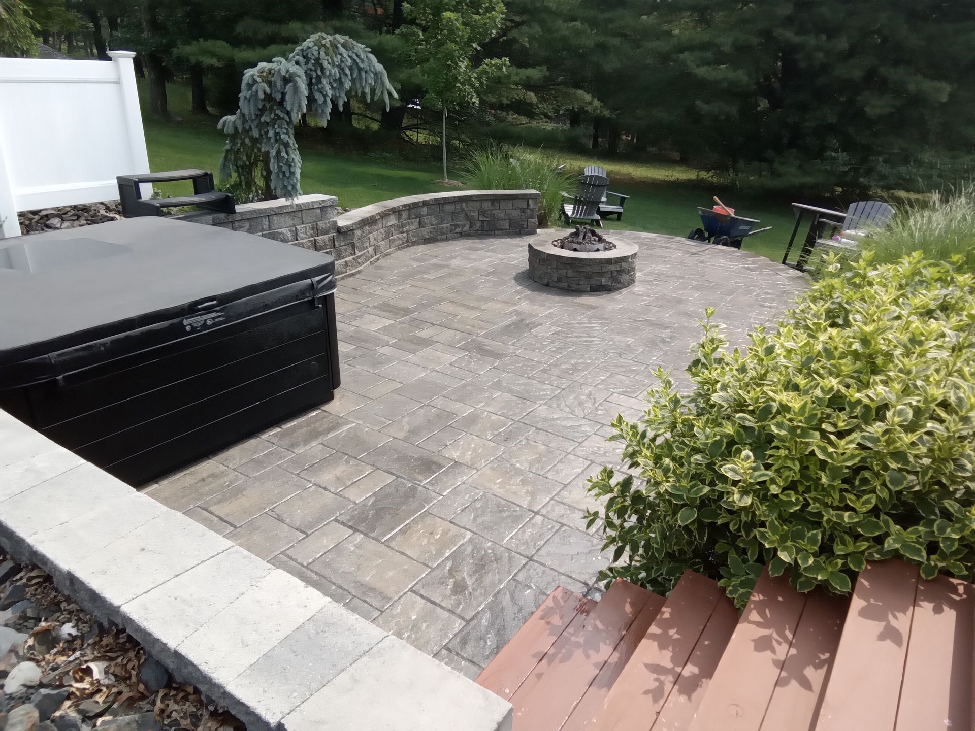 A paver patio with a fire pit, hot tub, and seating, bordered by a stone wall and landscaping.