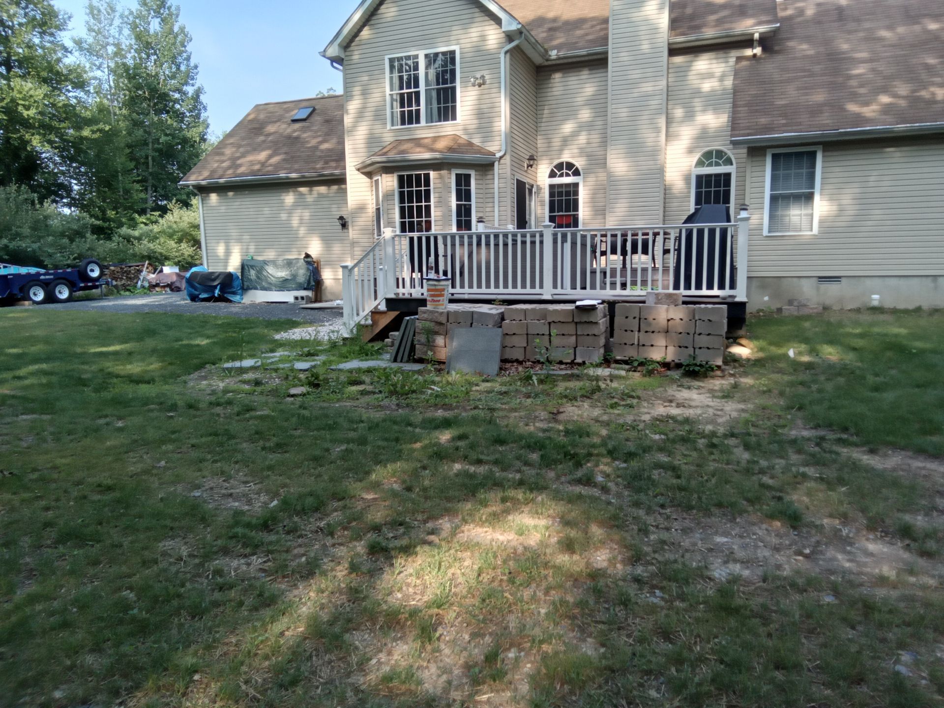 Backyard view of a house with a wooden deck, stacked paving stones, and patchy grass.