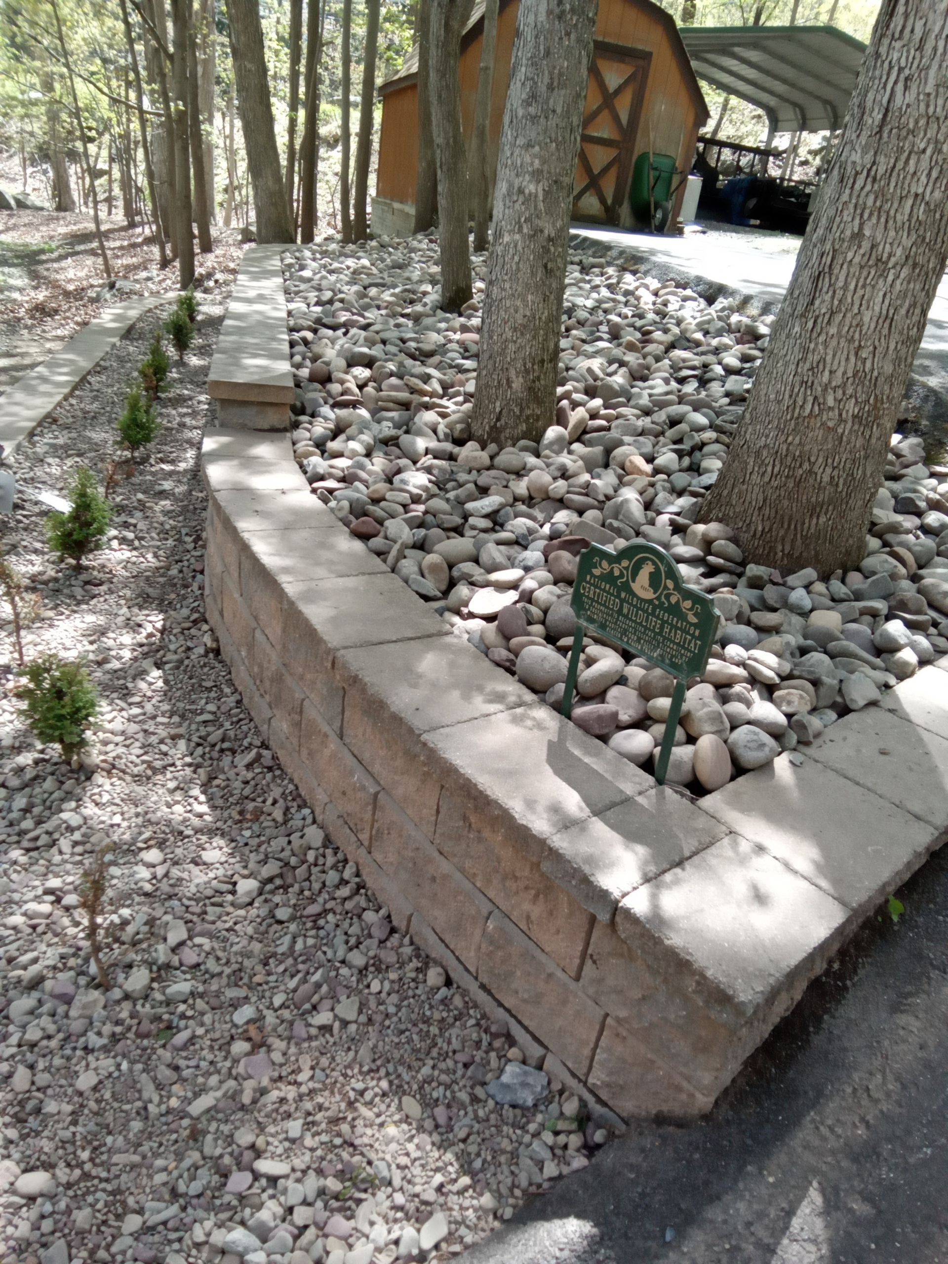 Stone retaining wall with trees, decorative rocks, and small shrubs.