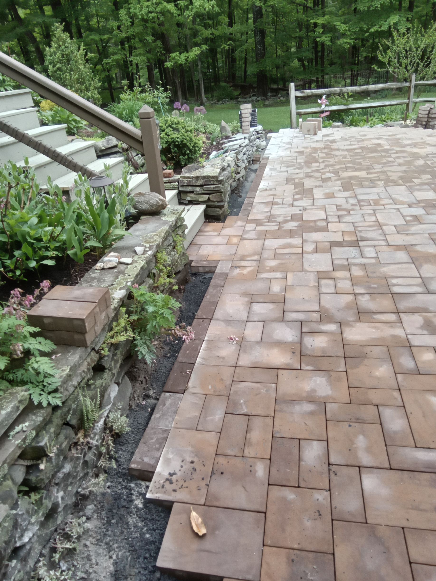 Stone patio with steps and landscaping, leading to a wooden fence and trees. Brown pavers.