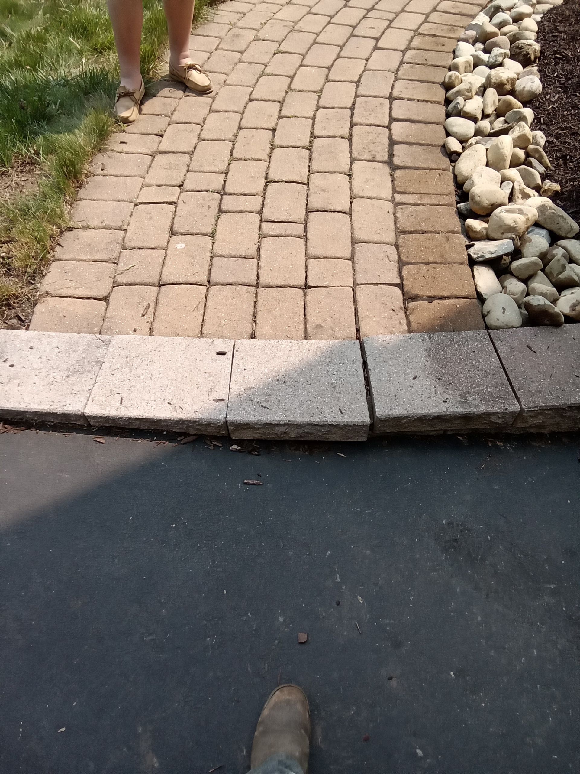 Brick pathway transitions to asphalt, person's feet visible, rocks border pathway.