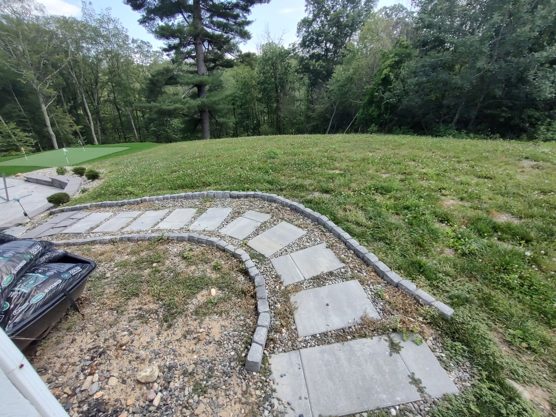 Stone path curves through a grassy yard leading to a small putting green, surrounded by trees.