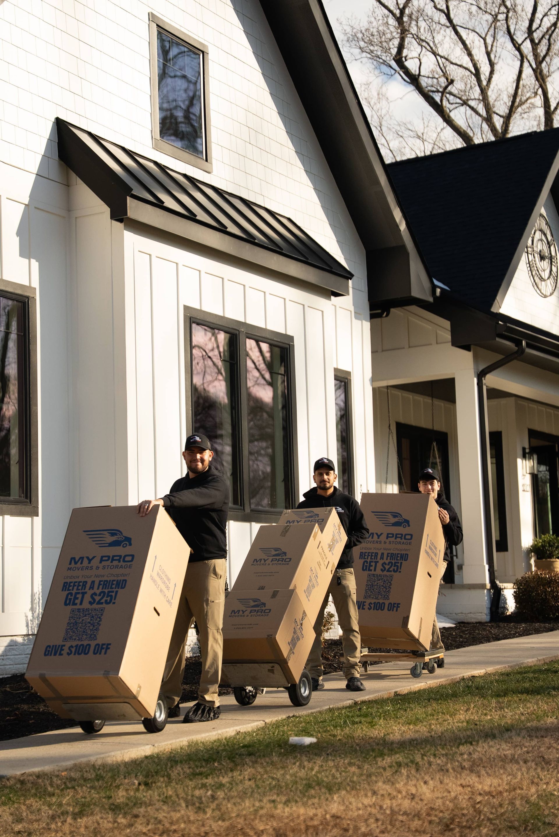 Three people carry moving boxes beside a white house on a sunny day.