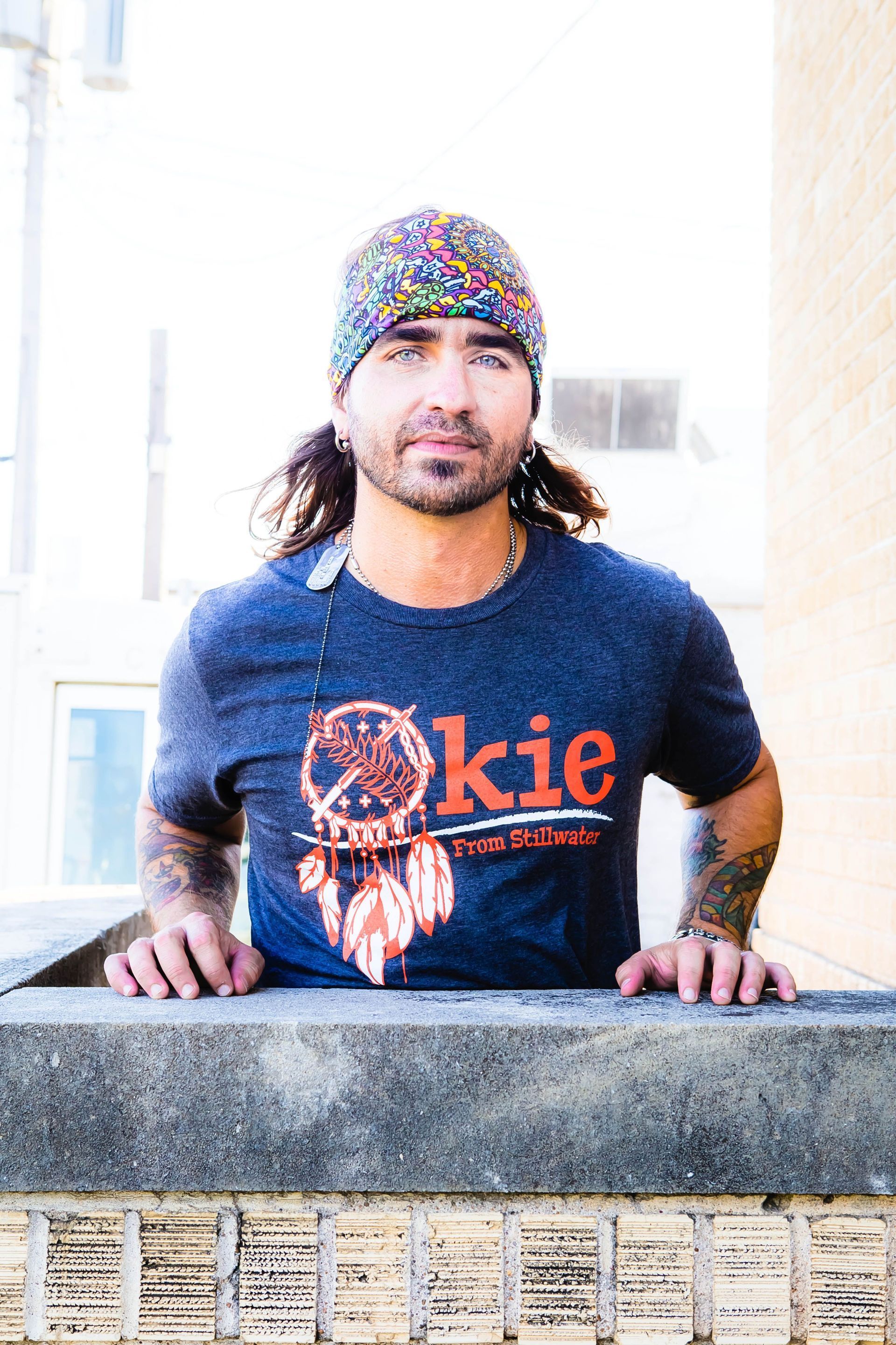 A man wearing a blue t-shirt and a beanie is leaning on a wall.