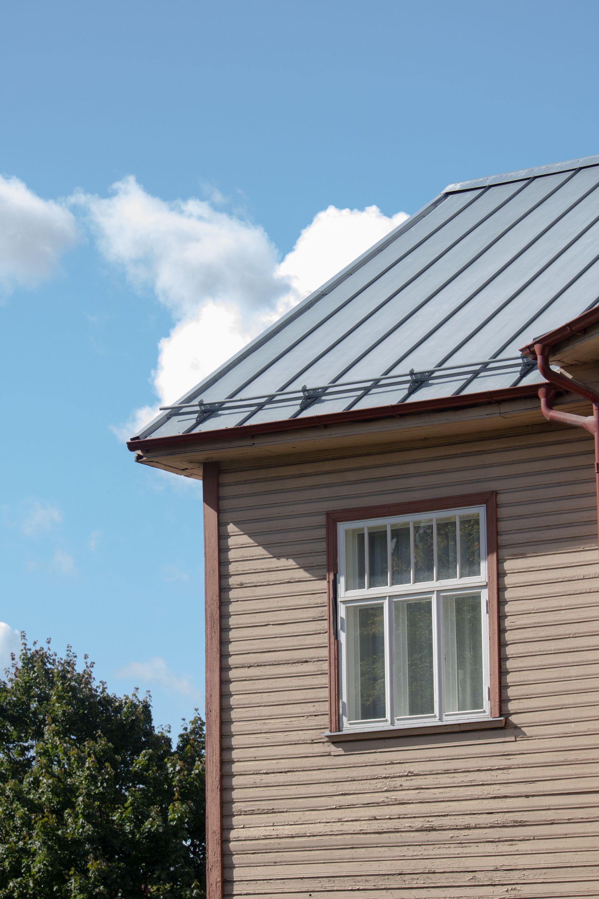 Corner of a house with a gray metal roof, beige brick wall, and white window against a blue sky.