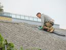 Worker kneeling on a steep roof, applying material with a trowel near a glass-railed building