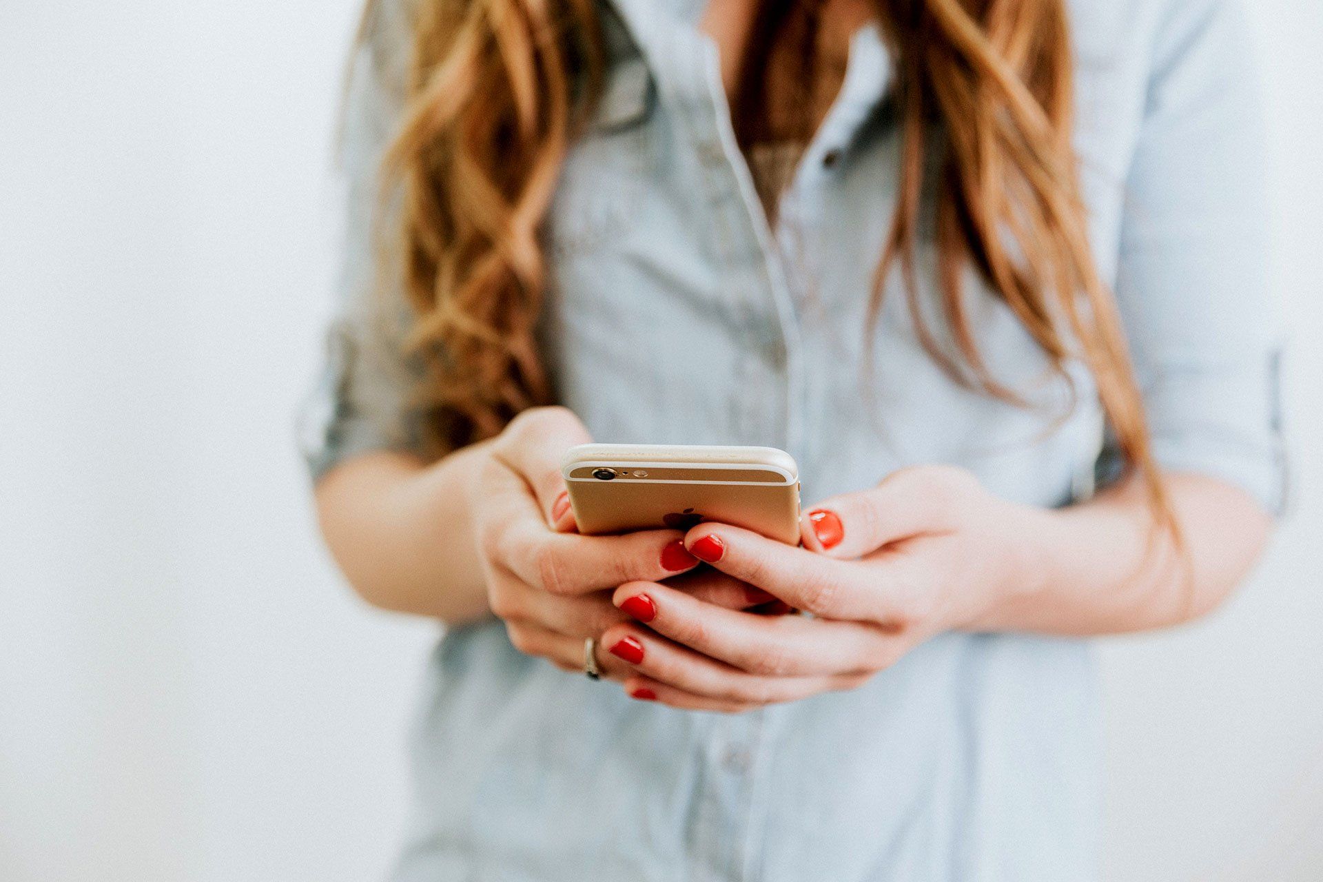 Woman in blue shirt holds a gold phone, hands with red nail polish. White background.