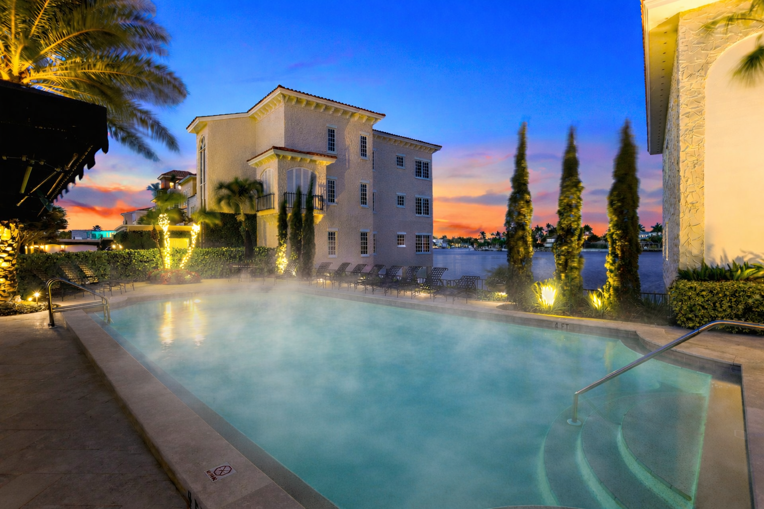 Outdoor swimming pool with steam, near a multi-story hotel at dusk; palm trees and lit walkways.