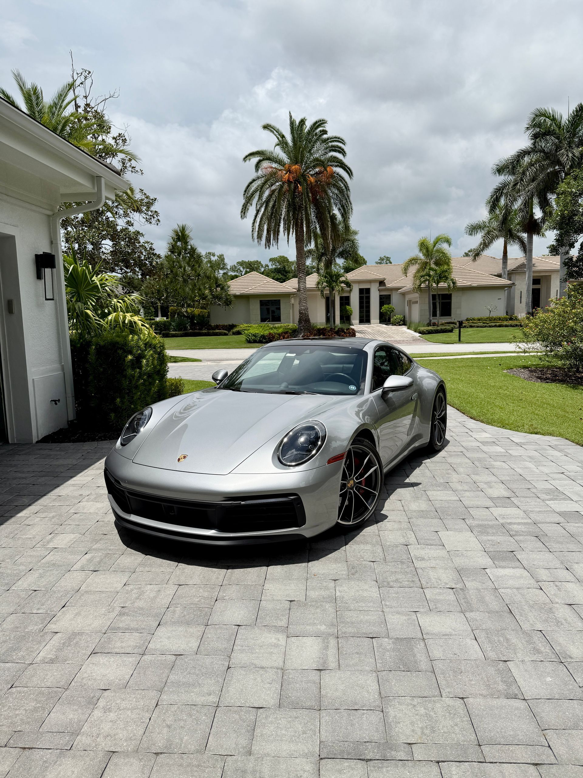 Silver Porsche 911 parked on a brick driveway in front of a house, palm trees in the background under a cloudy sky.