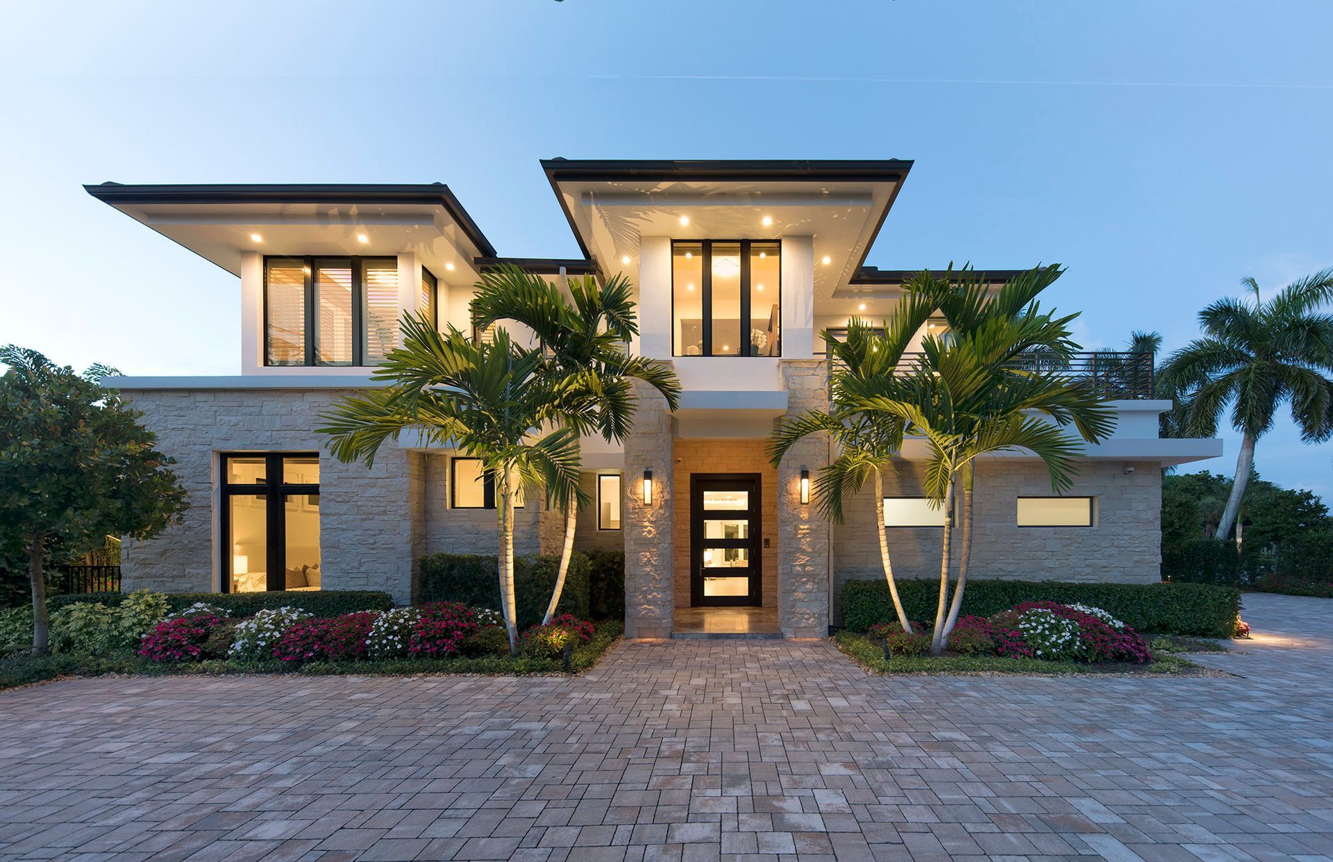 Modern two-story house with stone facade, palm trees, and illuminated windows at dusk.