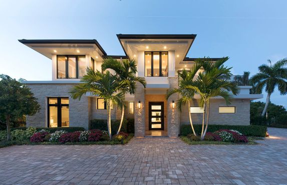 Modern two-story house with stone facade, palm trees, and illuminated windows at dusk.