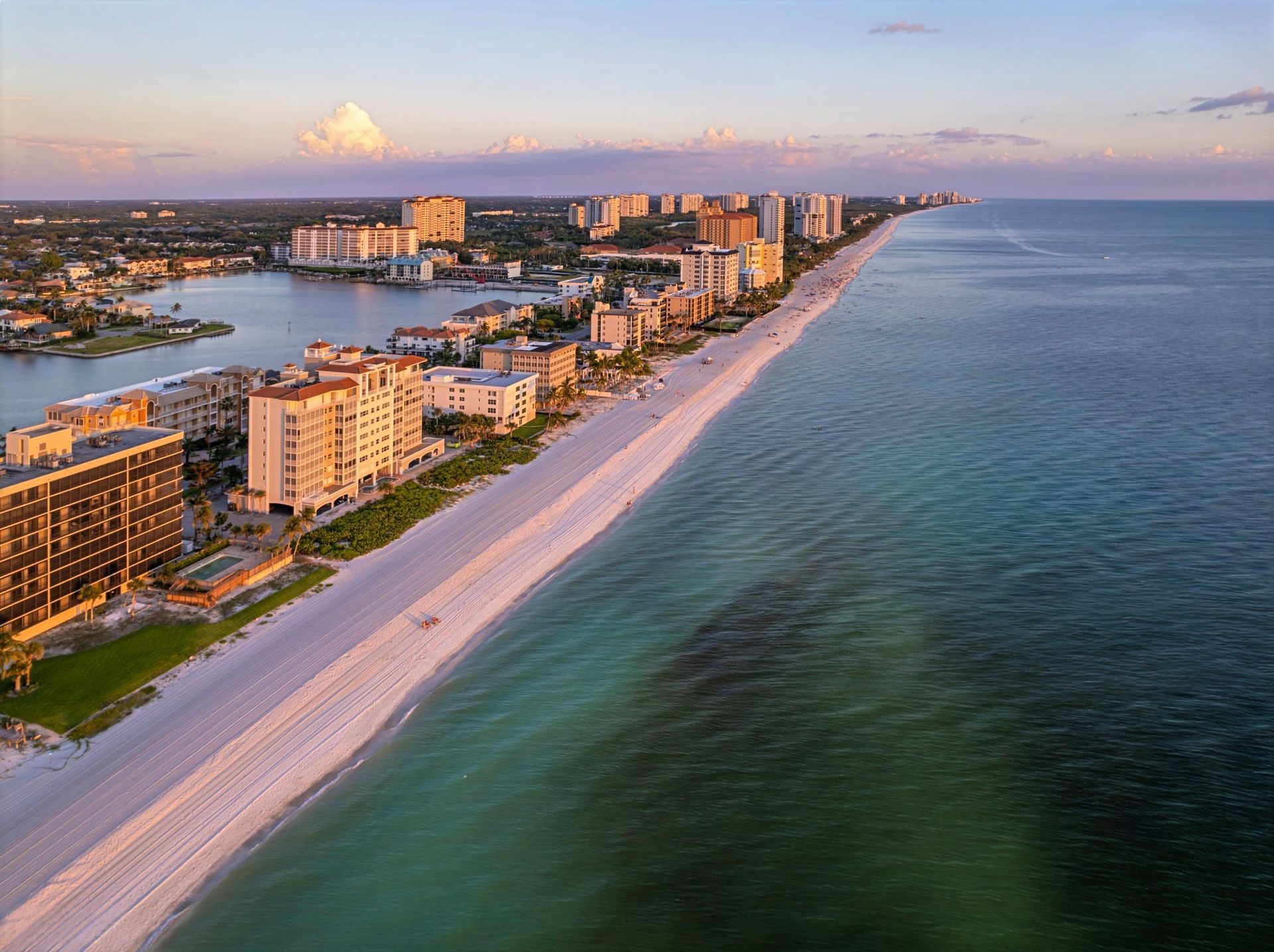 Coastal aerial view: Buildings line white sandy beach with turquoise water. Sunset colors in sky.