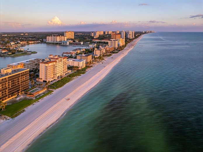 Coastal aerial view: Buildings line white sandy beach with turquoise water. Sunset colors in sky.