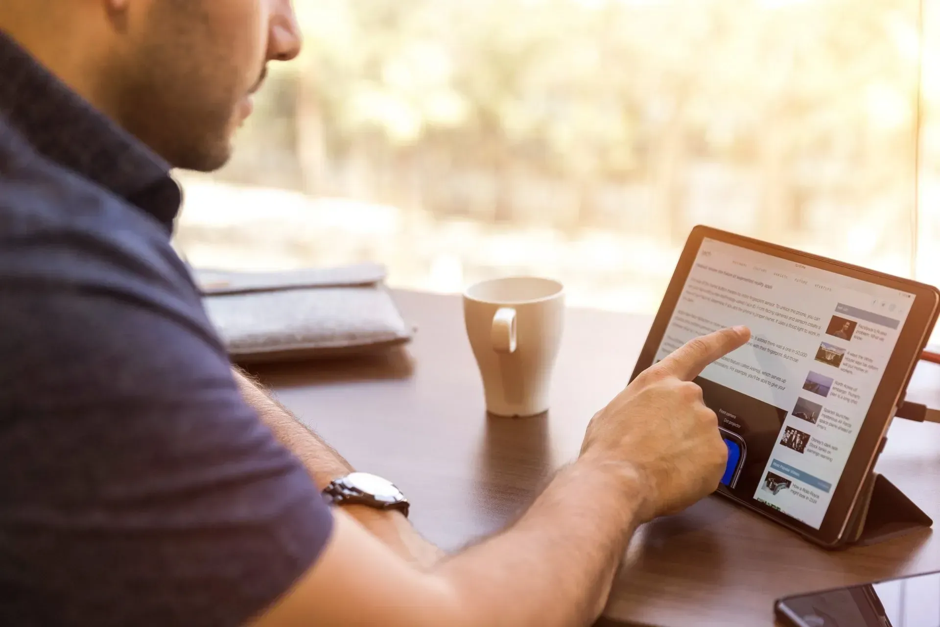 Man using a tablet at a table, tapping the screen with a finger. White mug and window in background.