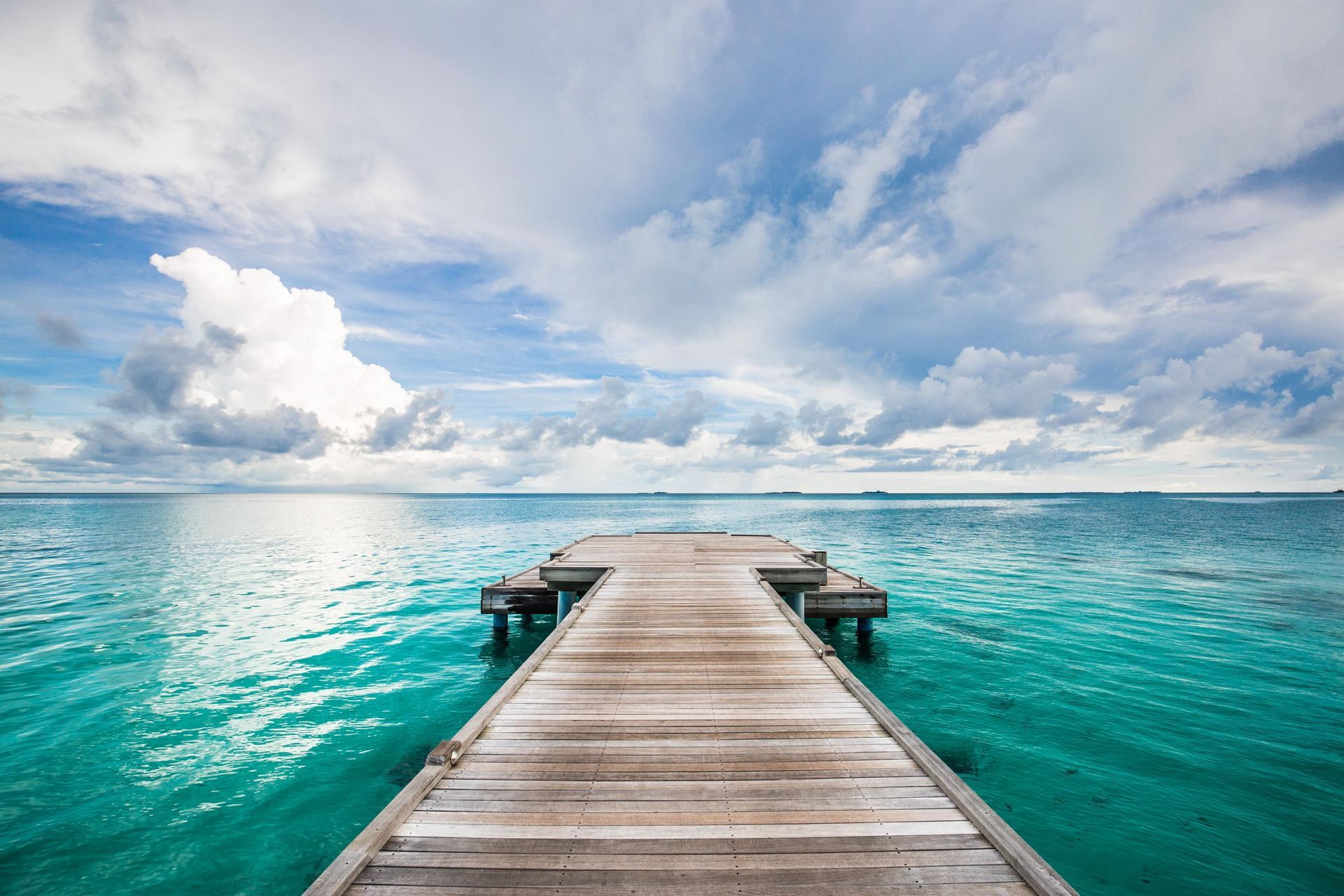 Wooden pier extends over turquoise water towards cloudy sky.