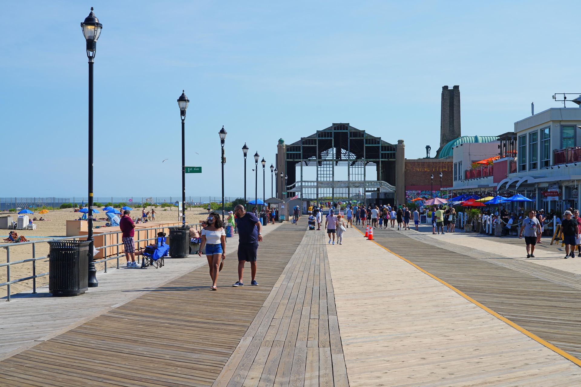 Boardwalk with people walking, beach on the left, shops and structures on the right, bright sunny day.