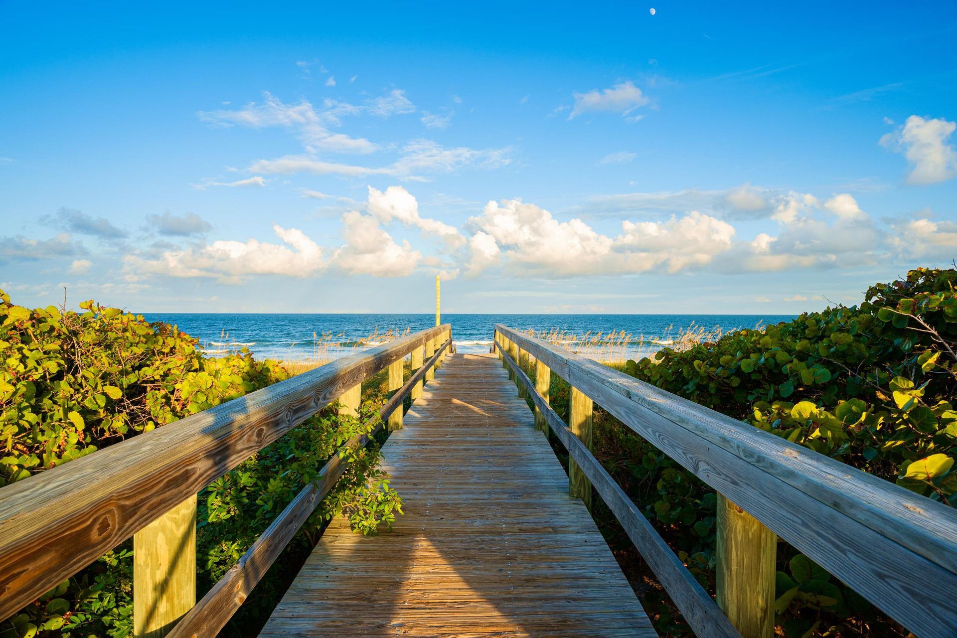 Wooden boardwalk leading to the ocean under a blue sky with fluffy clouds.