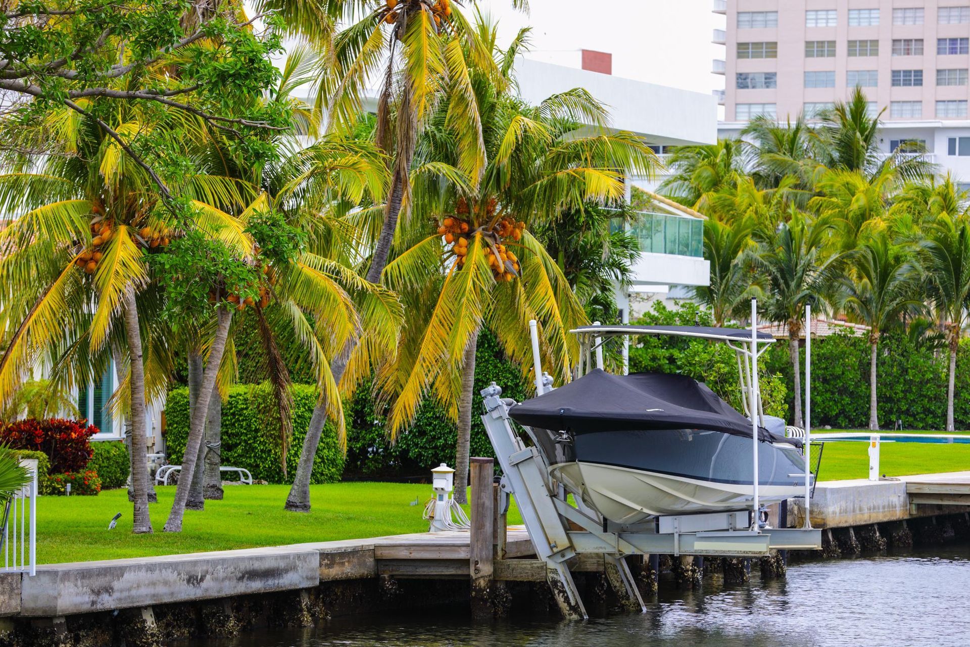 Boat lift with black cover, beside a green lawn with palm trees and a building in the background.