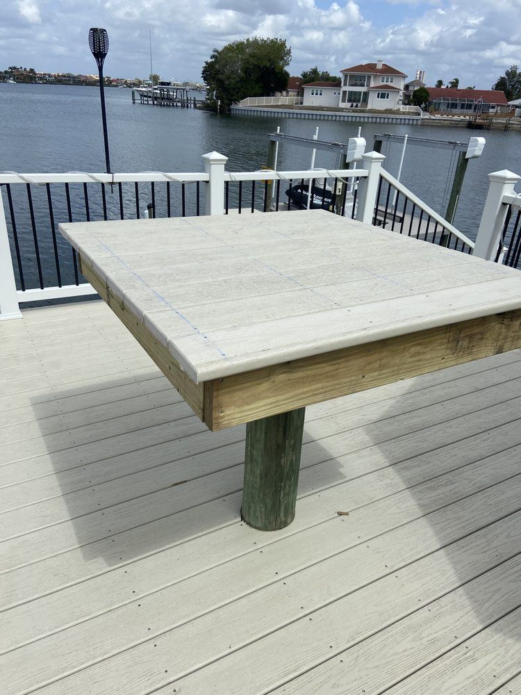 Square outdoor table on a wooden deck by water; light gray top, wooden base, railing, sunny day.