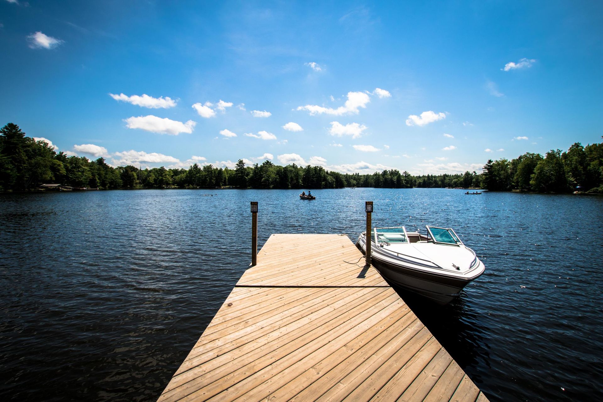 Wooden dock with a boat on a lake under a blue sky with fluffy clouds and trees.