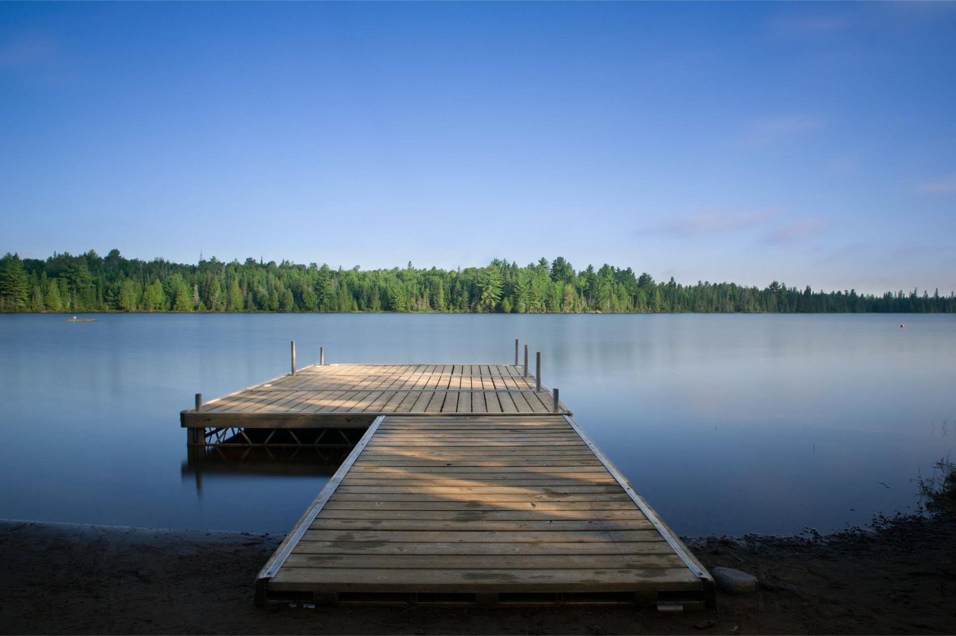 Wooden dock extending into a calm lake, surrounded by trees under a blue sky.