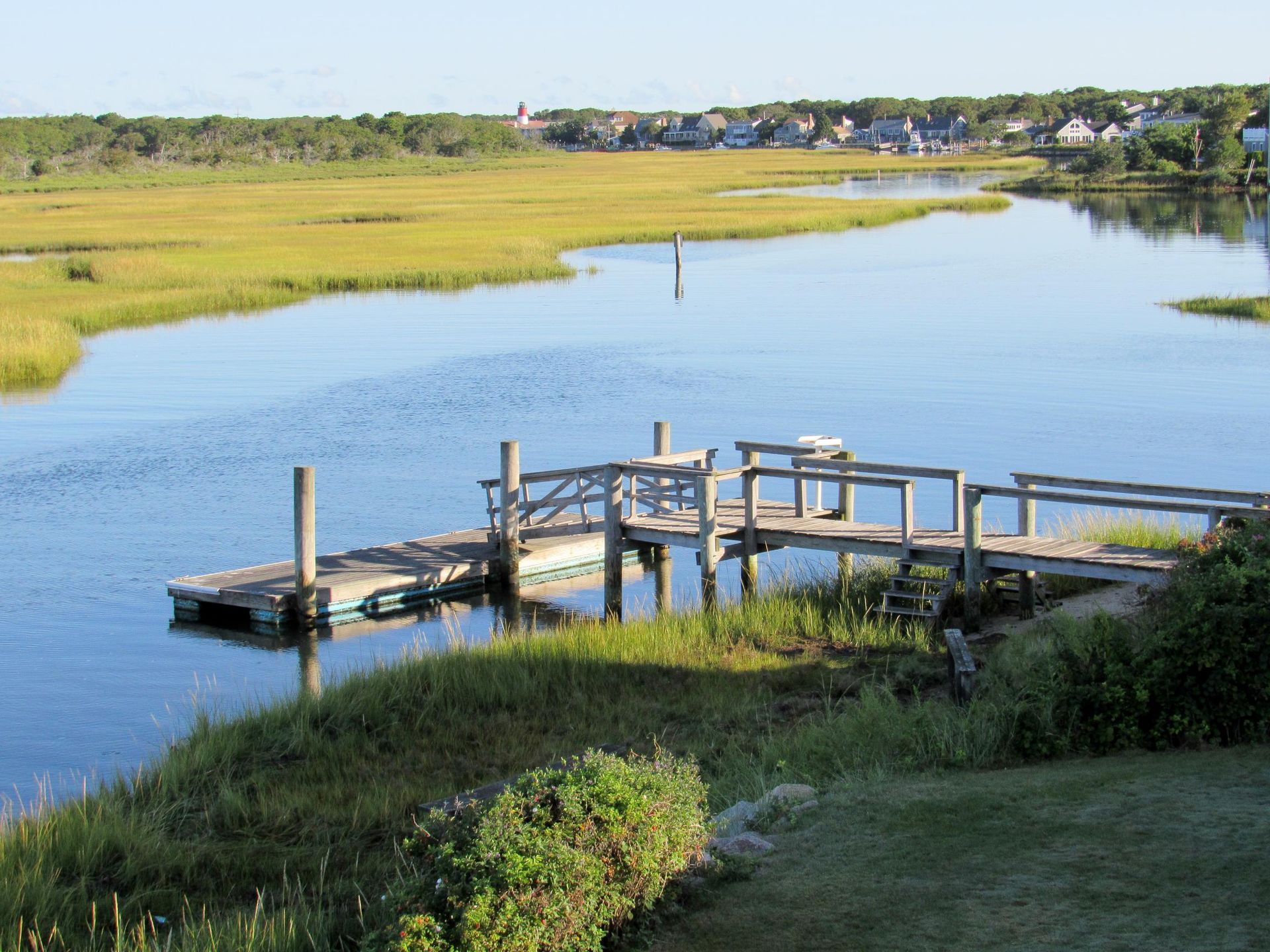 Wooden dock on a calm river surrounded by marshy grass and a distant town under a sunny sky.