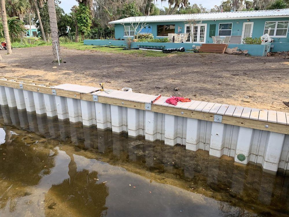 A newly constructed seawall borders a waterway, with a blue house in the background.