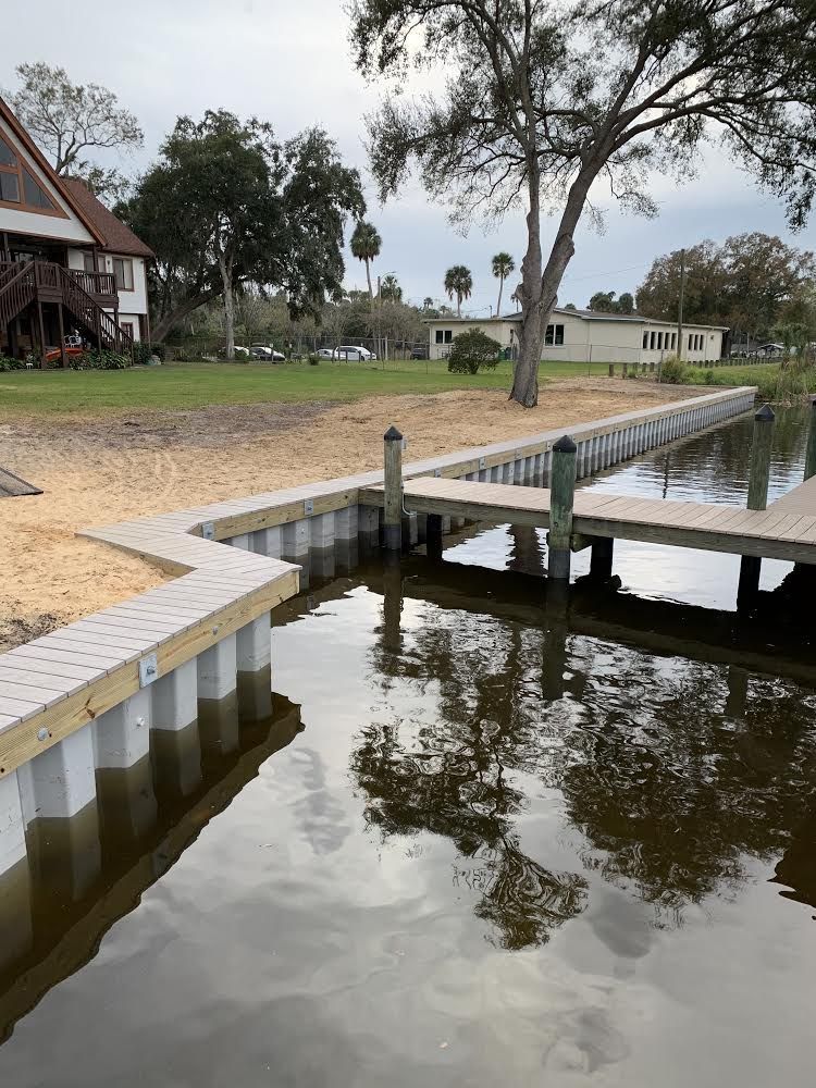 A dock with a sandy shore, set next to a calm body of water. Buildings and trees are in the background.
