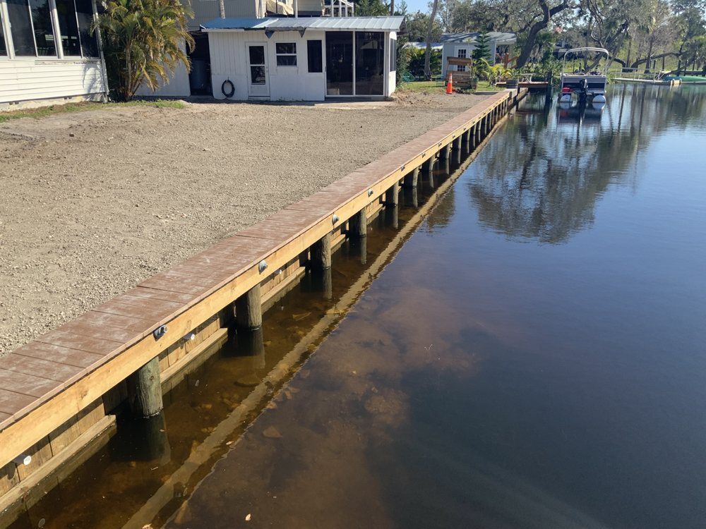Wooden dock extending into water next to a gravel area, with a small building in the background.