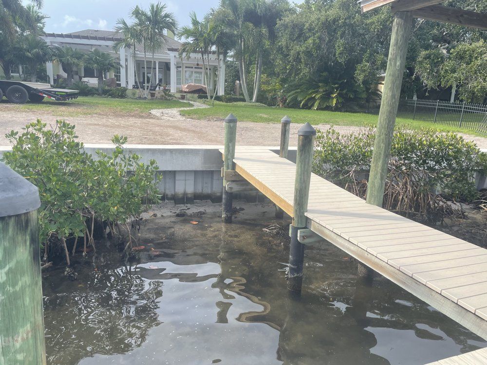 Dock with light-colored wooden planks extending from the shore into water, trees and a large house in the background.