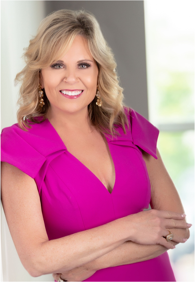 Lady Jen Du Plessis in a pink dress is standing with her arms crossed and smiling.