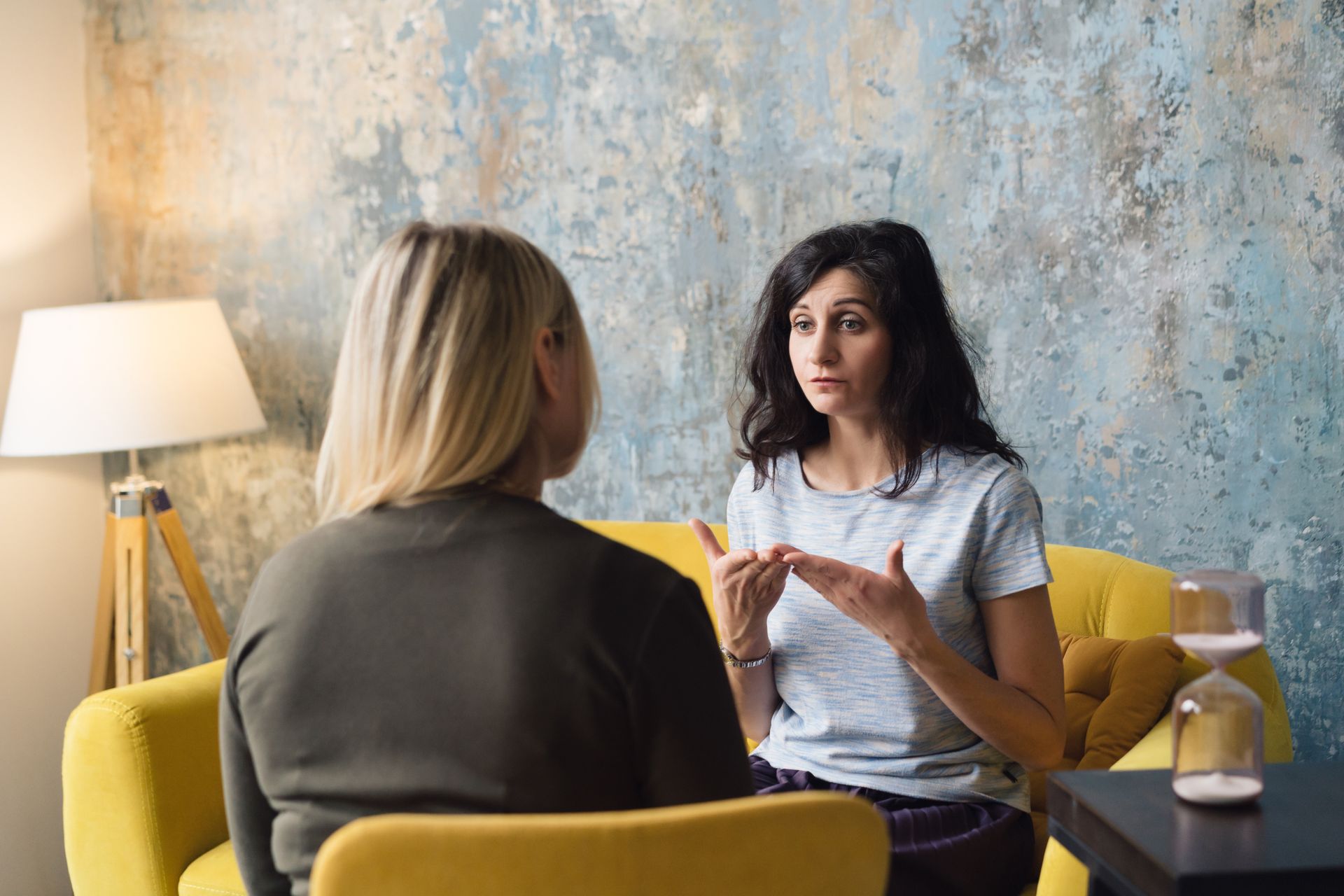 Two women are sitting on a yellow couch talking to each other.