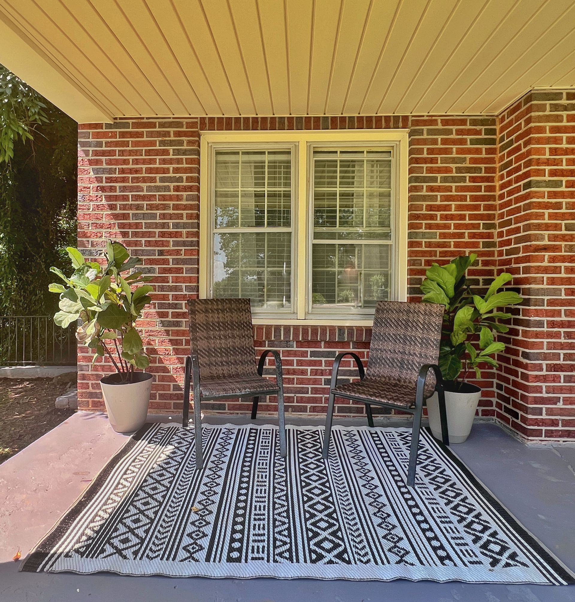 Front porch with chairs on a patterned rug, flanked by potted plants, beneath a window and brick facade. 