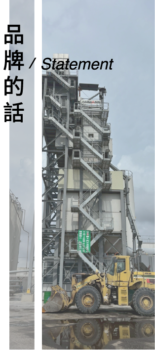 在陰沉的天空下，一輛黃色前裝式裝載機停在一座高大的灰色工業混凝土或瀝青攪拌站前。