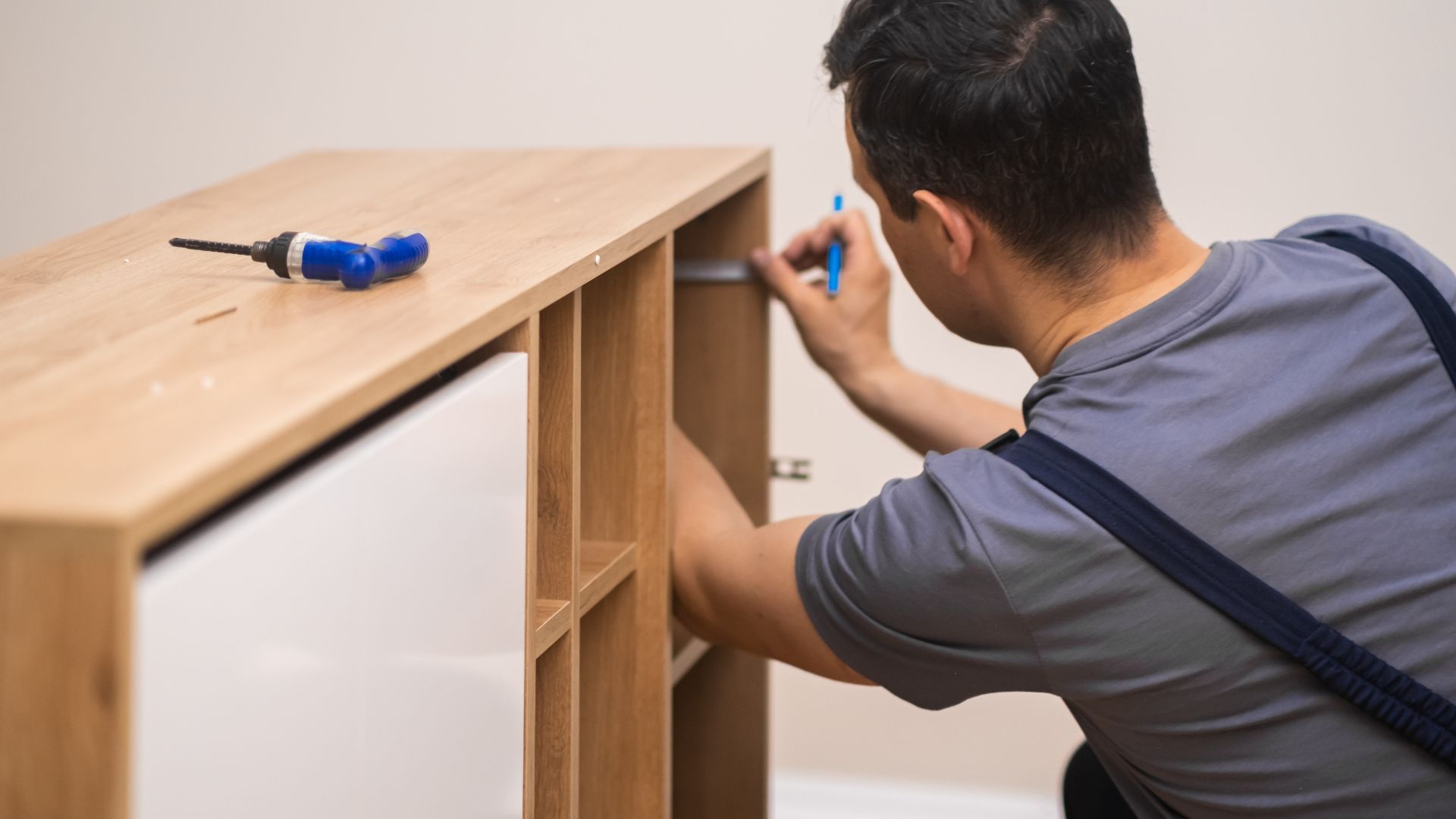 A worker in a gray uniform assembling a light wood shelving unit with a blue tool nearby.