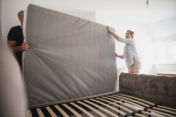 Two people moving a large gray mattress into a bedroom with a bed frame