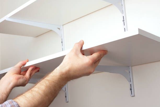 Hands installing a white shelving board onto wall-mounted metal shelf brackets.