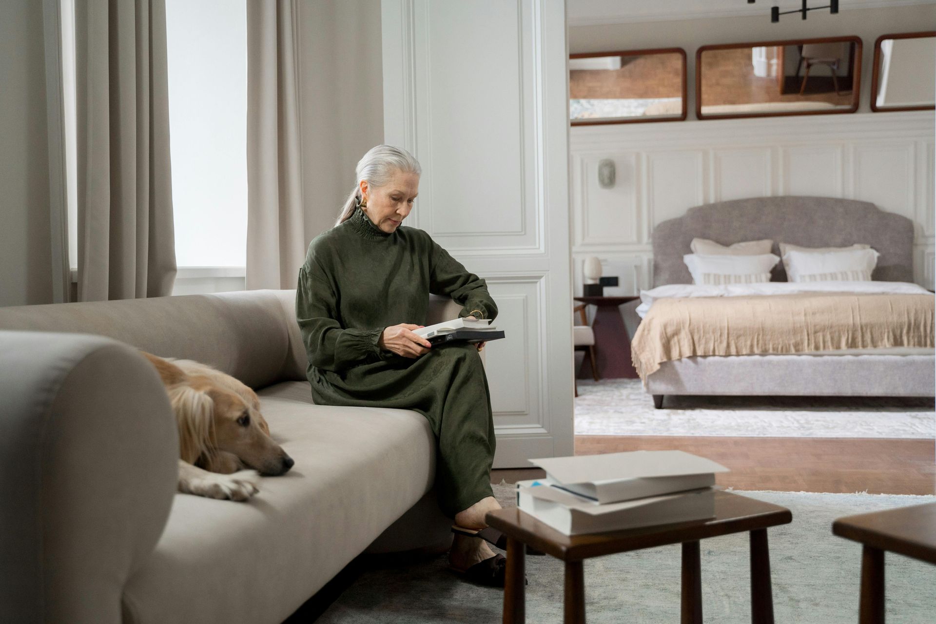 A person sits on a light gray sofa with a dog, holding a tablet, with a bedroom visible in the background.
