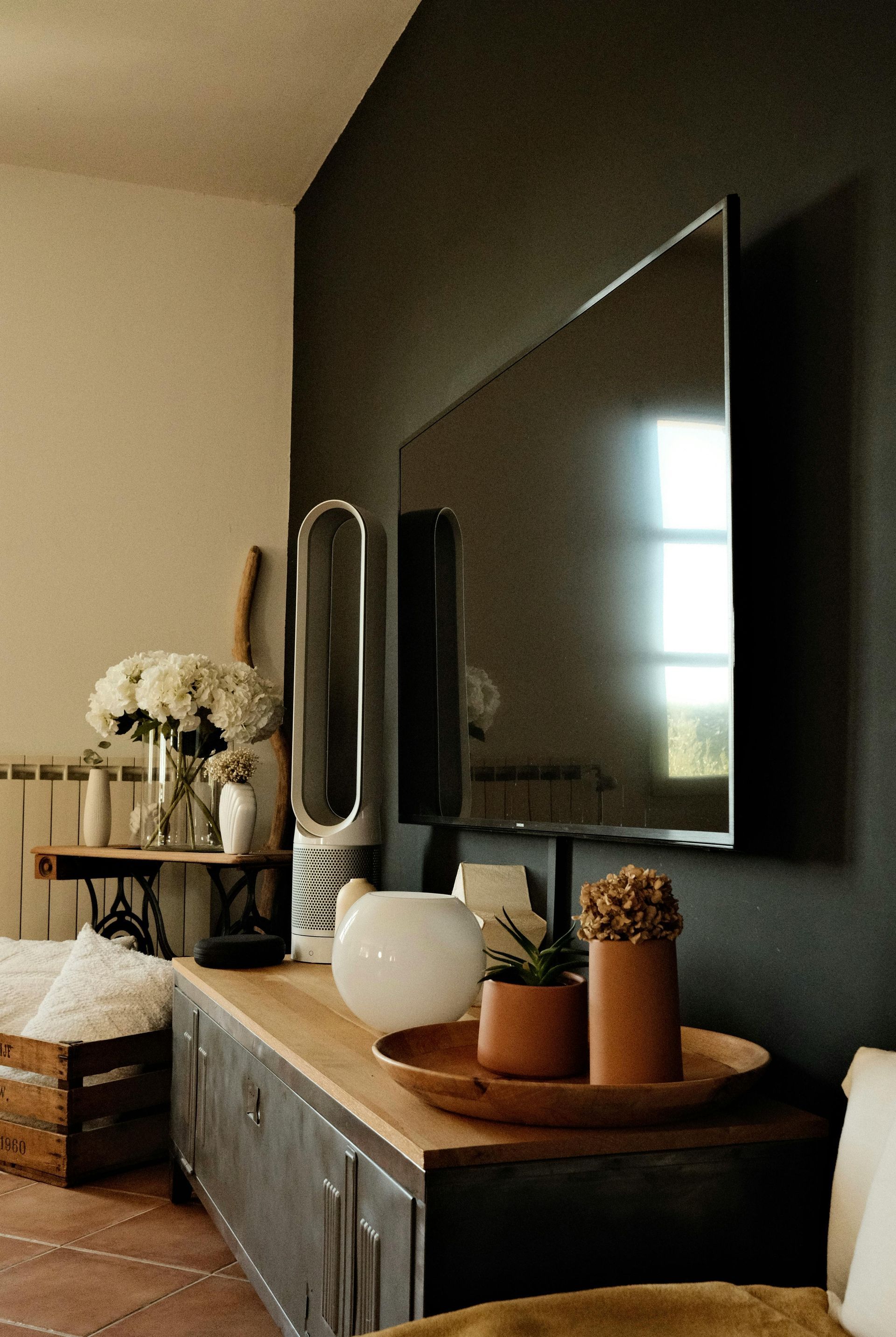 A TV mounted on a dark wall above a wooden console table featuring a Dyson fan, potted plants, and a vase of flowers.