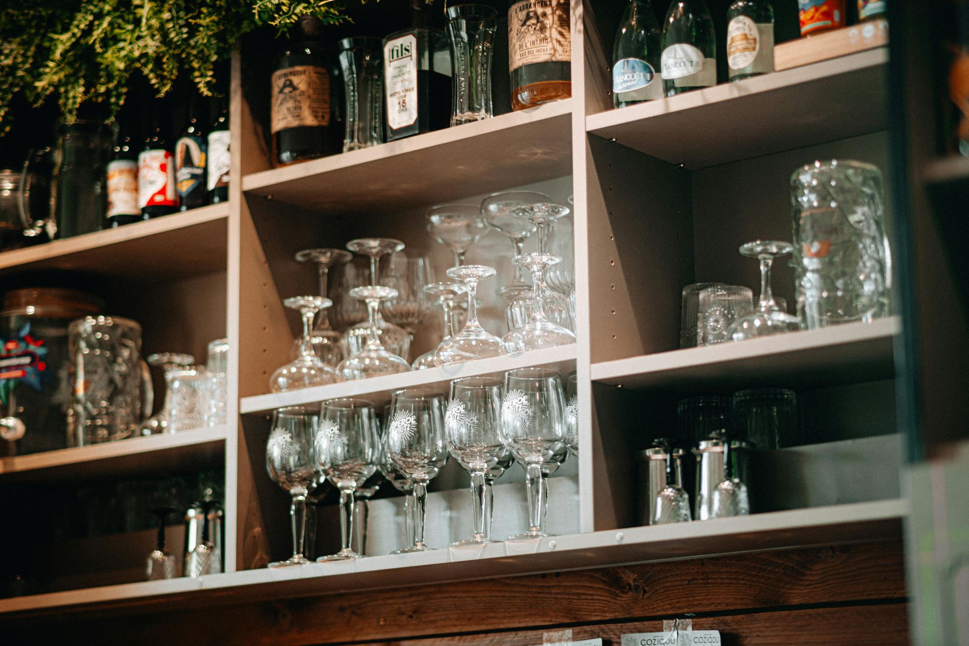Wooden shelves in a bar stocked with various glass bottles, wine glasses, and mugs against a dark background.